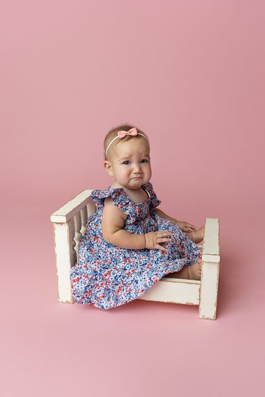 Baby girl in a floral dress sitting on a white bench during a studio milestone portrait
